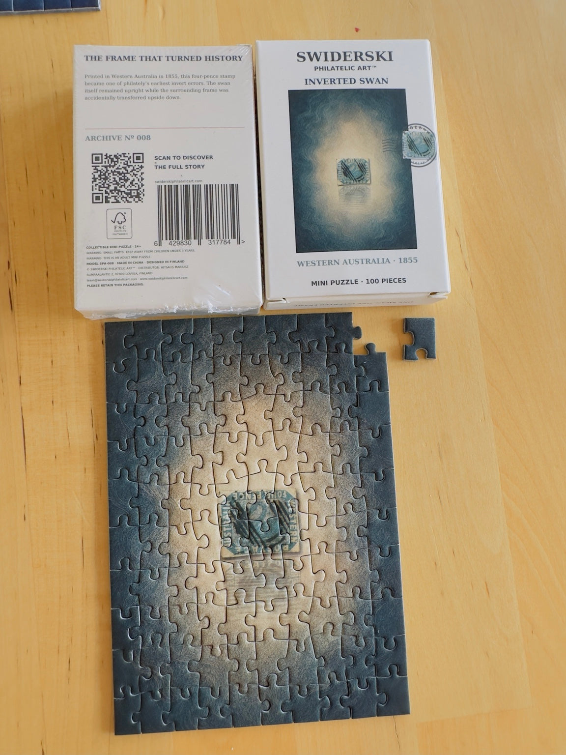 Inverted Swan mini puzzle almost assembled, showing a loose piece and the front and back of its packaging.